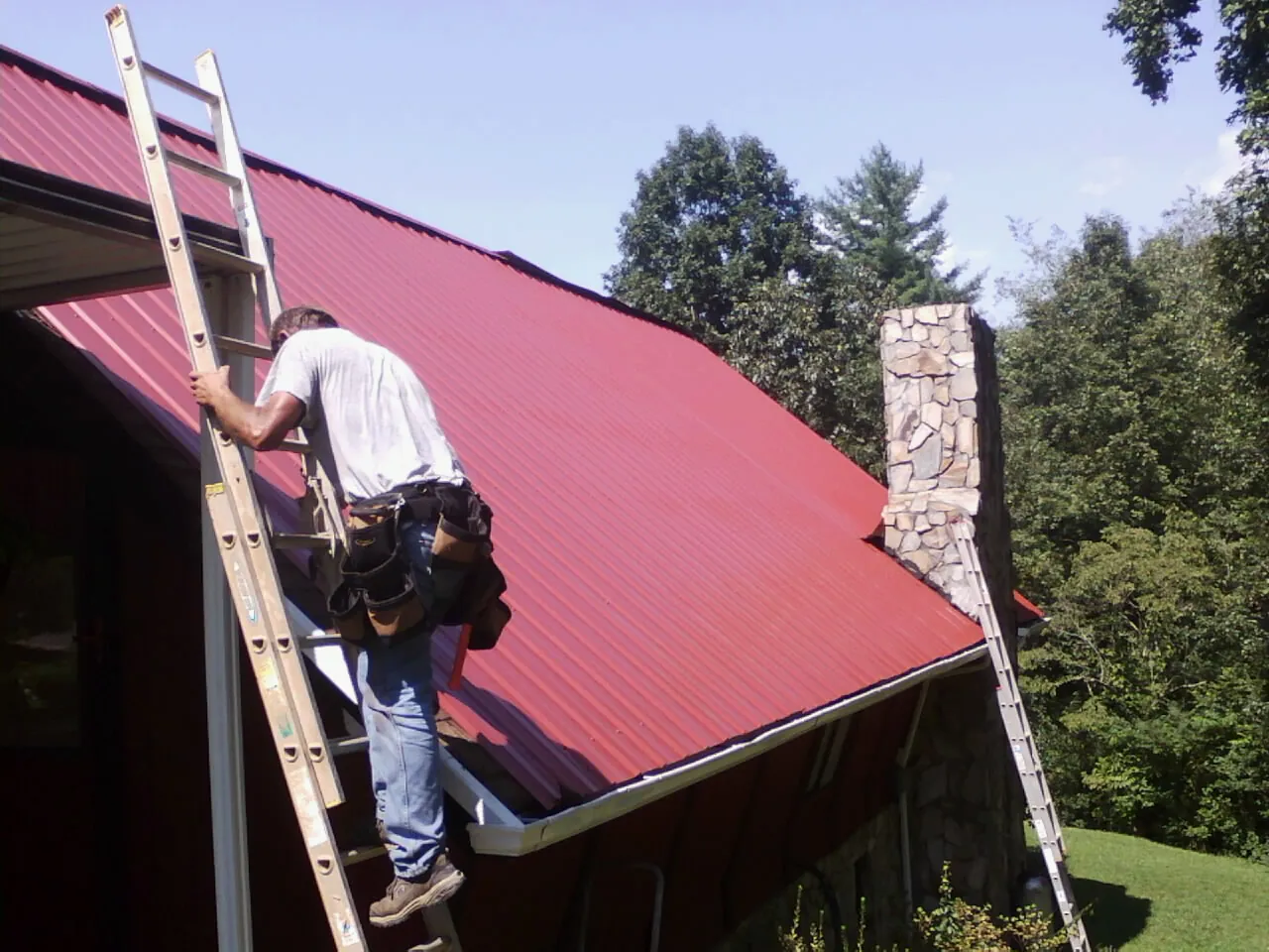 Roofer on a ladder beside a red metal roof and stone chimney — Soffit & Fascia Repair by Roofing Brothers in Janesville